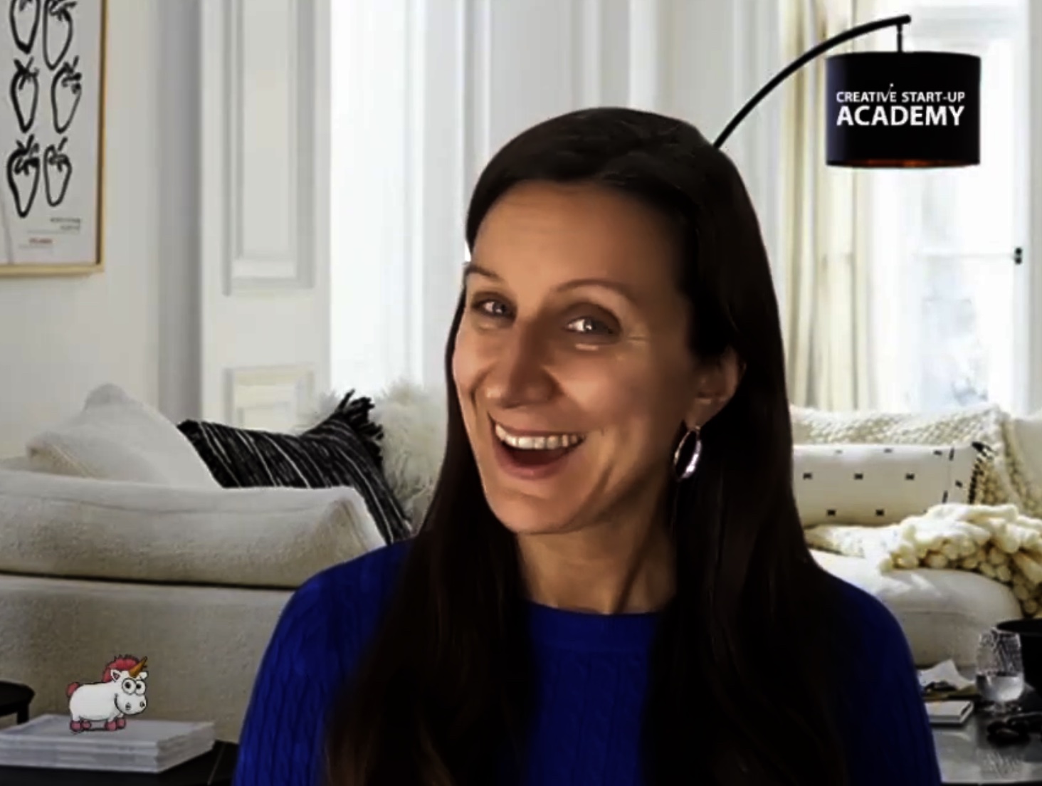 A woman with long brown hair and a blue sweater is smiling in a cozy living room with white walls, a beige sofa, and a lamp. The image includes a "Creative Start-Up Academy" logo in the upper right corner.